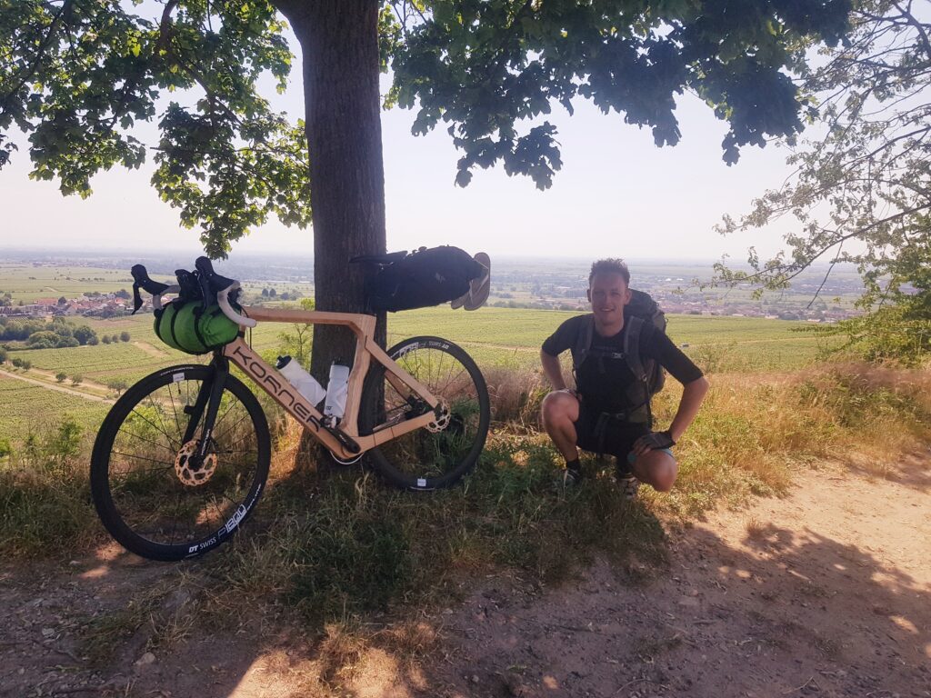 The wooden road bicycle standing to a tree. Sjors posing crouched right to it. Photo taken from a hill and so with a view on the terrain on the back