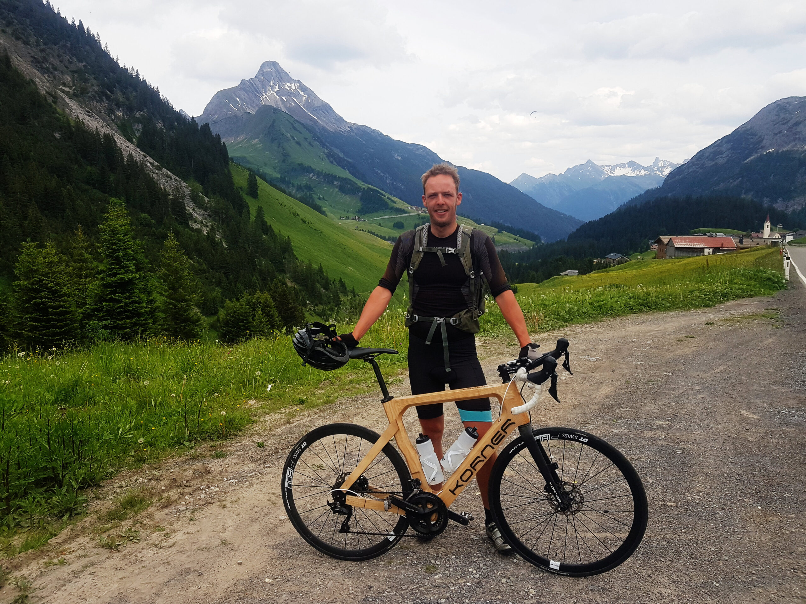 Sjors on the side of the road with his wooden bicycle in his hands on a gravel surface. On the background you see the Austrian Alps.